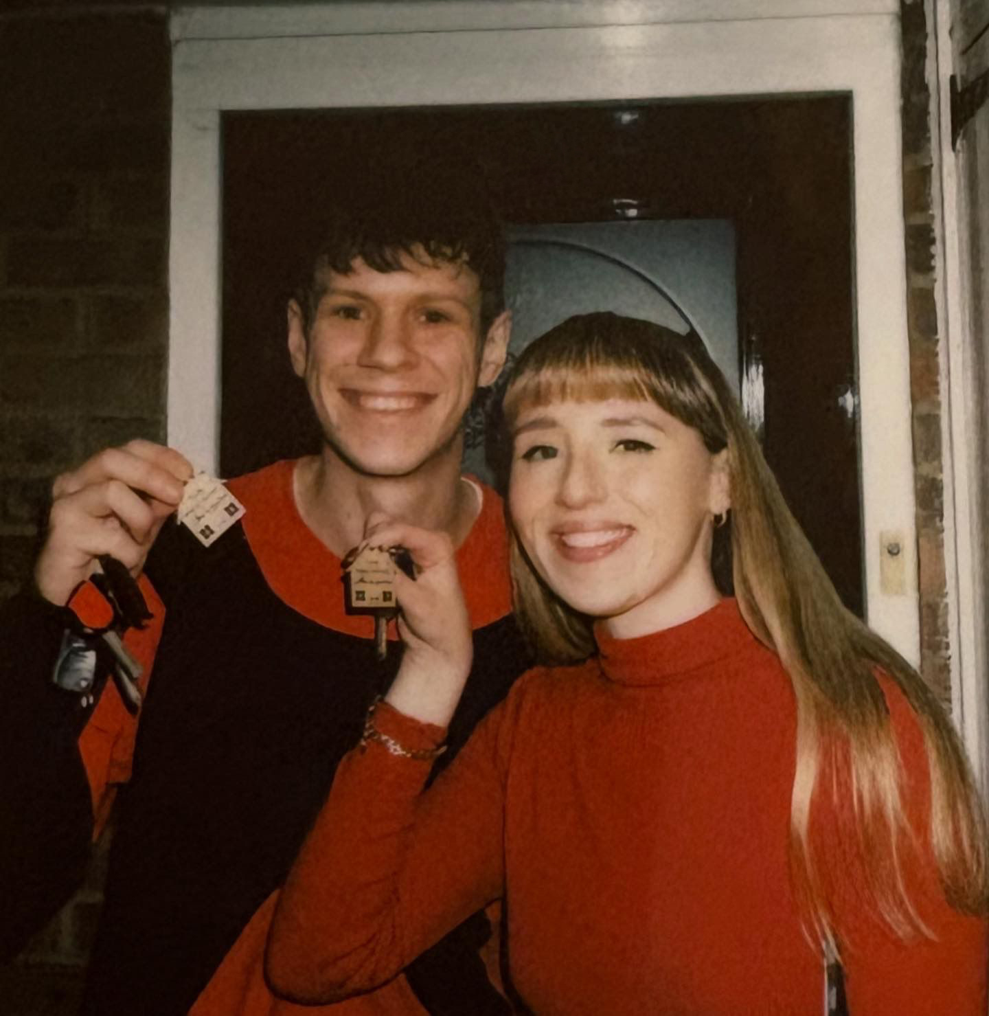 Alex and Lauren smiling at the front door of their first home, each holding a set of keys with wooden house keyrings